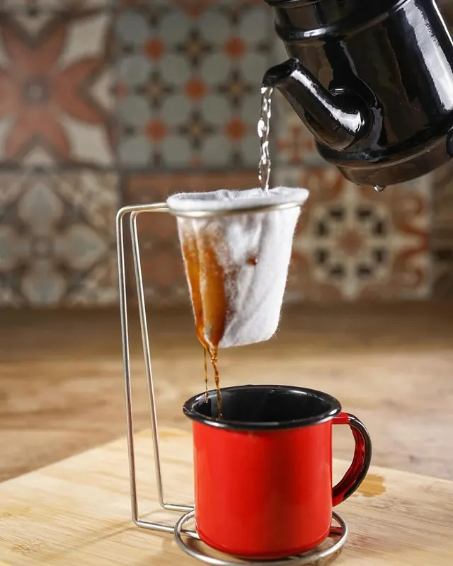 Café sendo coado no pano, ao lado de uma caneca esmaltada vermelha, com ladrilho hidráulico ao fundo