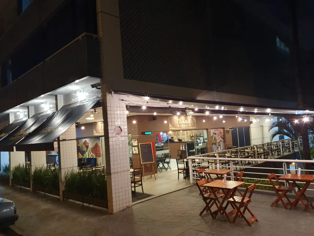Frente do restaurante à noite, com varal de luzes, mesas de madeira na calçada e o letreiro Minas Bistrô
