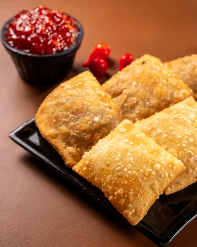 Pastéis fritos numa travessa preta, com geleia de pimenta num potinho e pimentas vermelhas ao lado